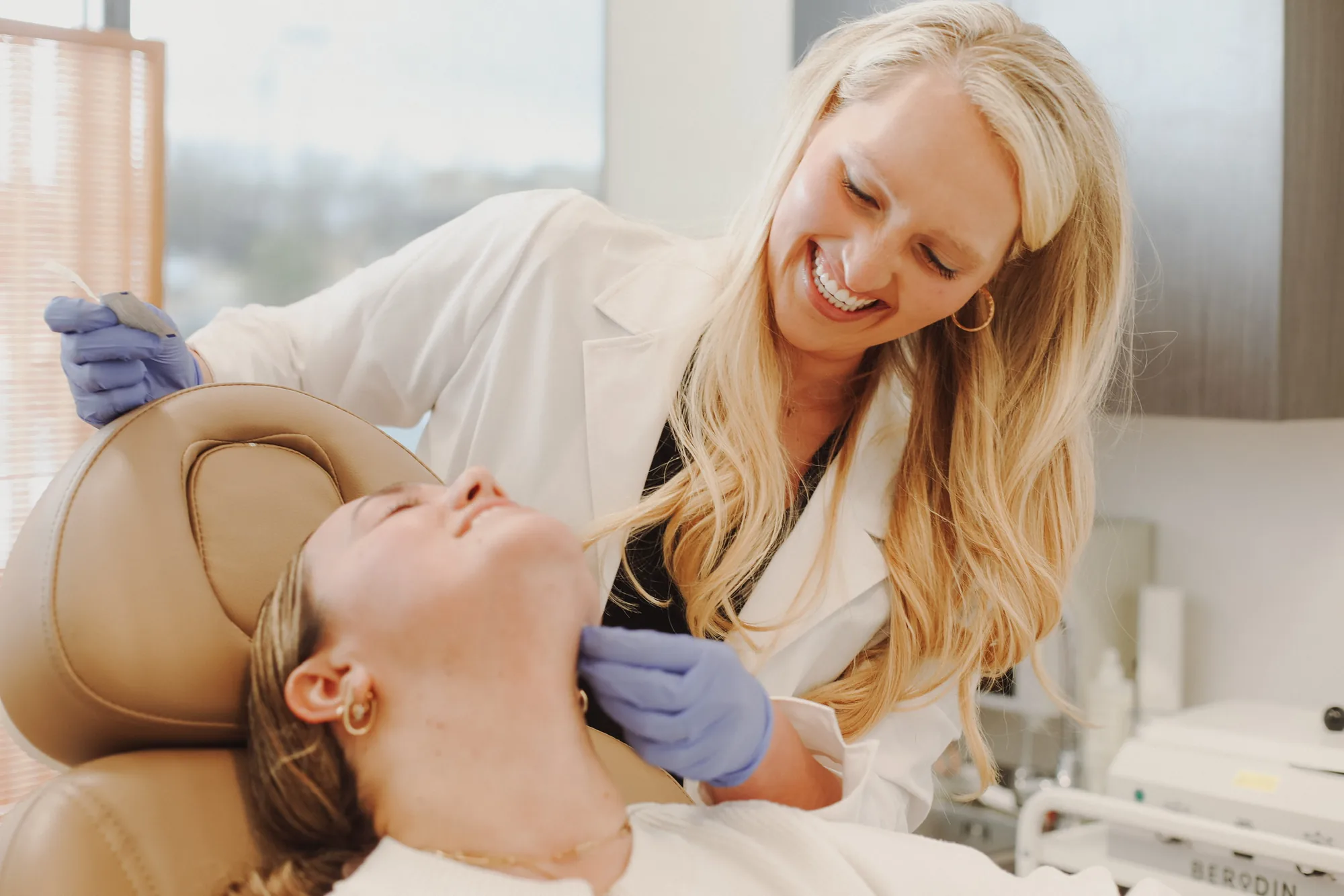 Provider in a white coat gently examining a reclined client's neck during a skin treatment