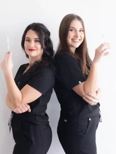 Two clinicians in black scrubs standing back to back, each holding a syringe.